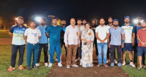 रात्रि क्रिकेट टूर्नामेंट के दौरान मैदान में खड़े अतिथि संध्या डालाकोटी, किरण डालाकोटी और दोनों टीमों के खिलाड़ी, रोशनी में खींची गई सामूहिक फोटो।